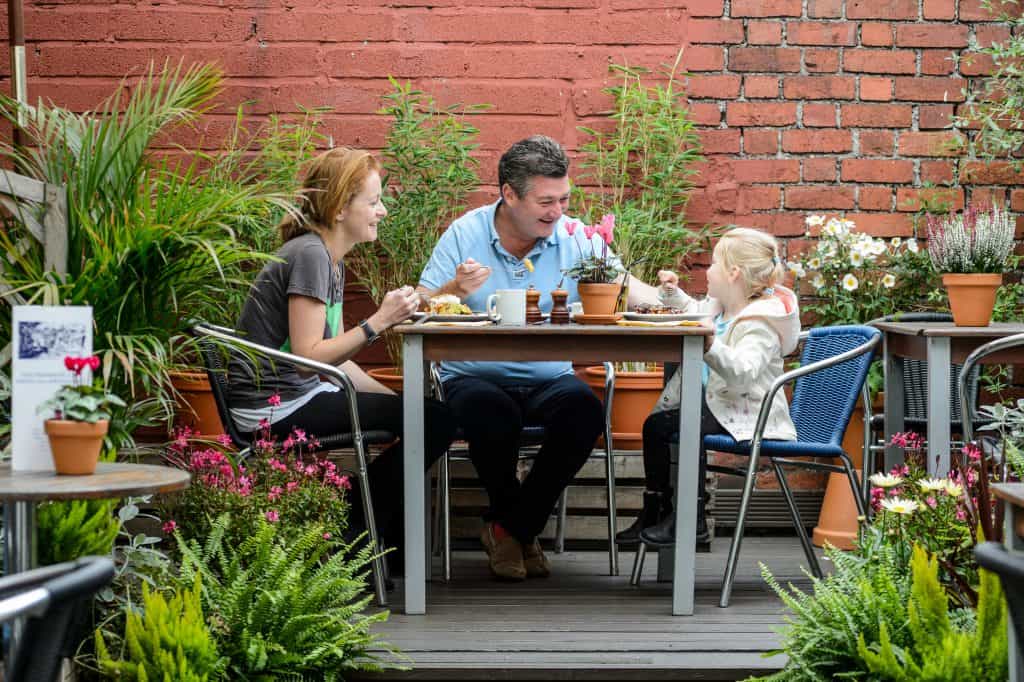 Families seated at outdoor tables against a red brick wall and greenery at Primrose Cafe