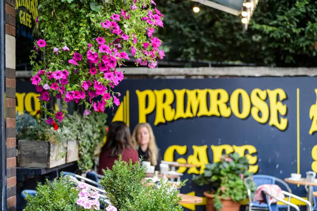 Primrose Cafe frontage on a sunny day, with hanging flowers, the blue Primrose Cafe sign, and pavement seating in Clifton Village, Bristol
